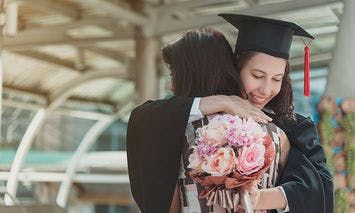 A graduate in a cap and gown embraces another person while holding a bouquet of pink flowers, standing under a glass-roofed structure in an outdoor setting.