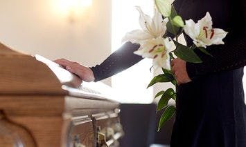 A person dressed in black rests a hand on a wooden casket and holds white lilies, standing in a dimly lit room with soft light filtering through.