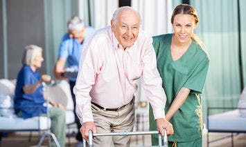 An elderly man uses a walker with assistance from a caregiver in green scrubs, set in an outdoor elderly care facility where other seniors interact in the background.